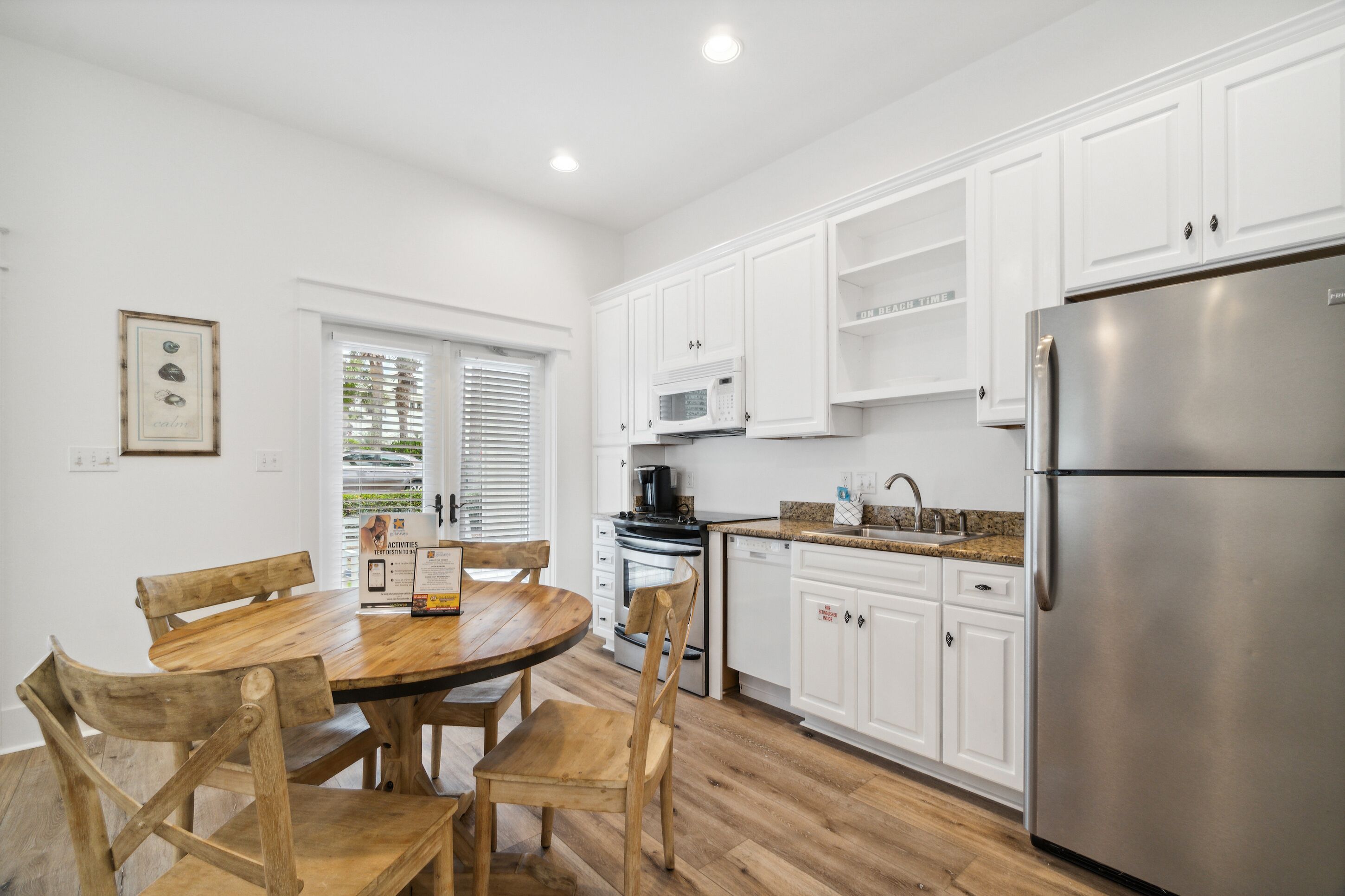 A modern kitchen with white cabinets, a stainless steel refrigerator, a wooden dining table, and a window with blinds.