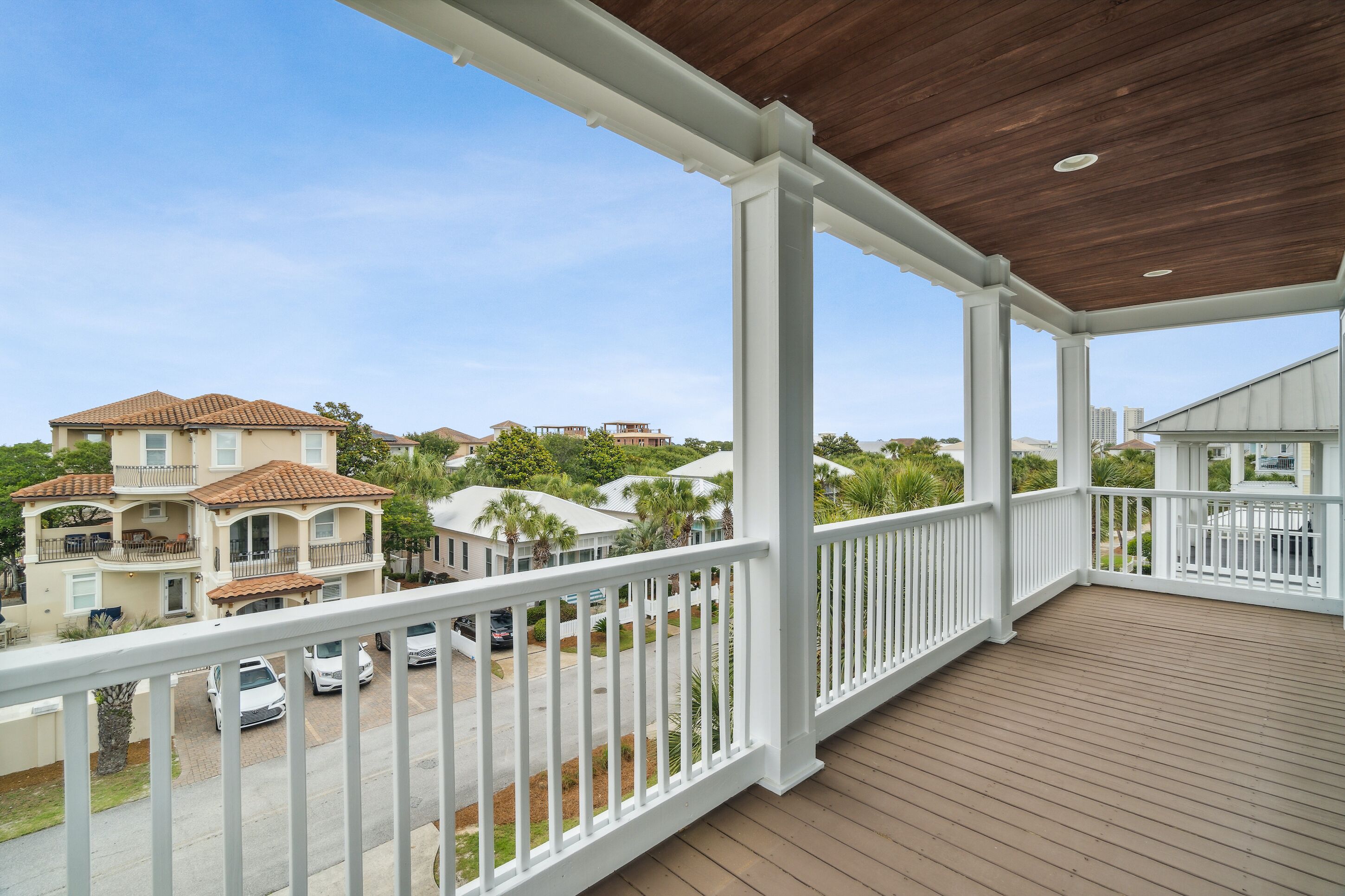 A covered balcony with white railings and a wooden floor, overlooking a residential area with houses and parked cars.