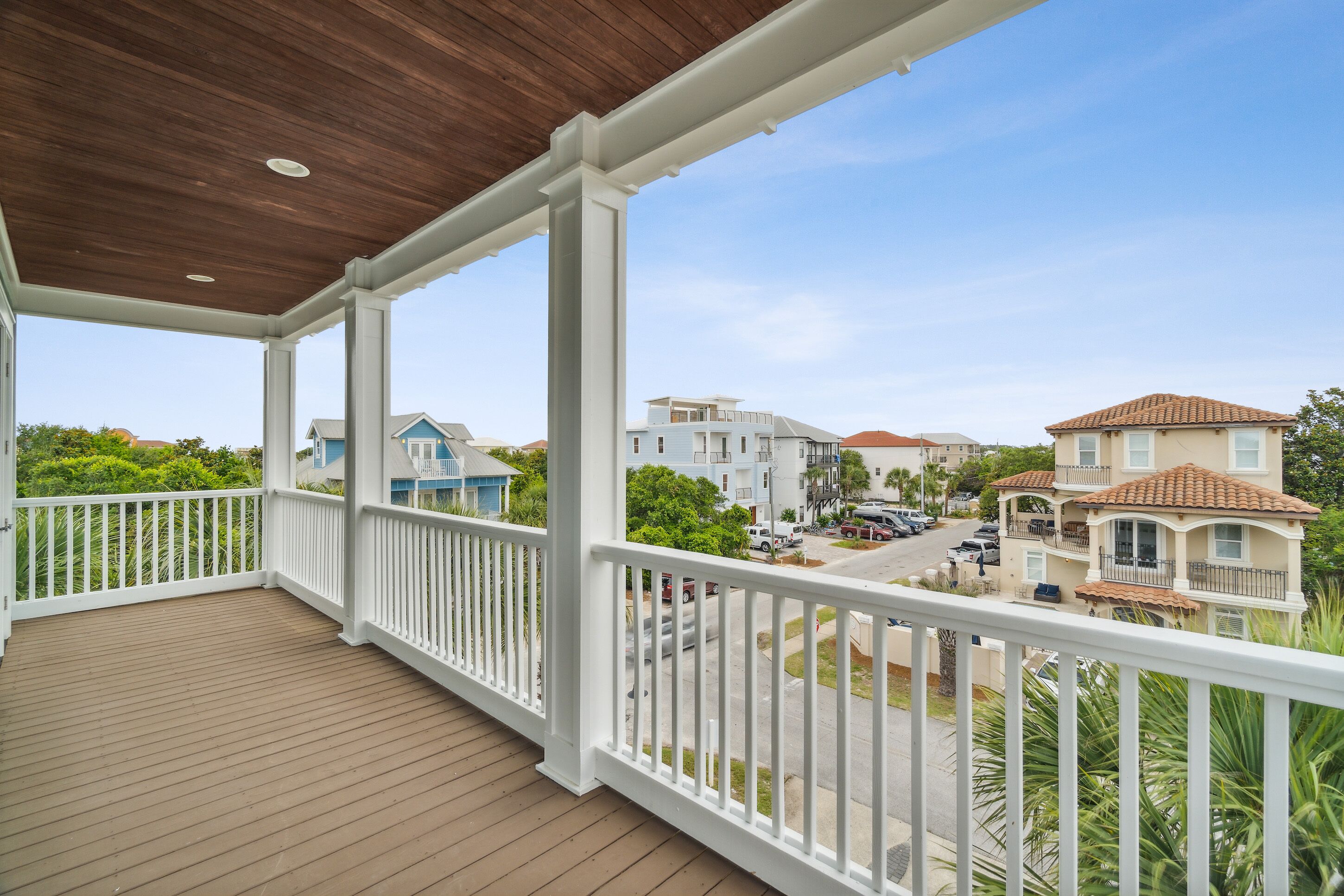 A covered balcony with a wooden floor and white railings, overlooking a residential street with houses and parked cars.