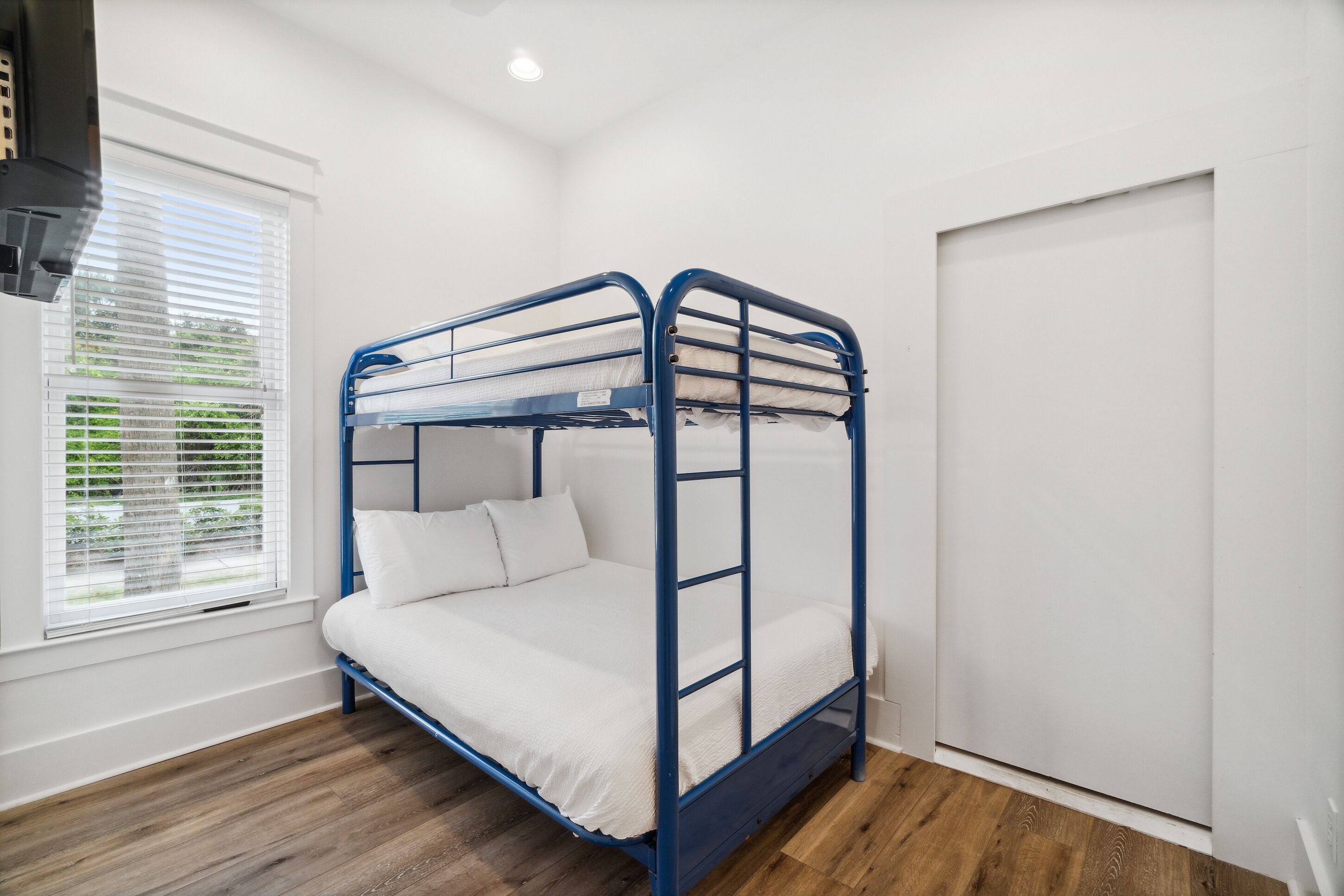 A bunk bed with white bedding in a room with wooden flooring and a window with blinds.