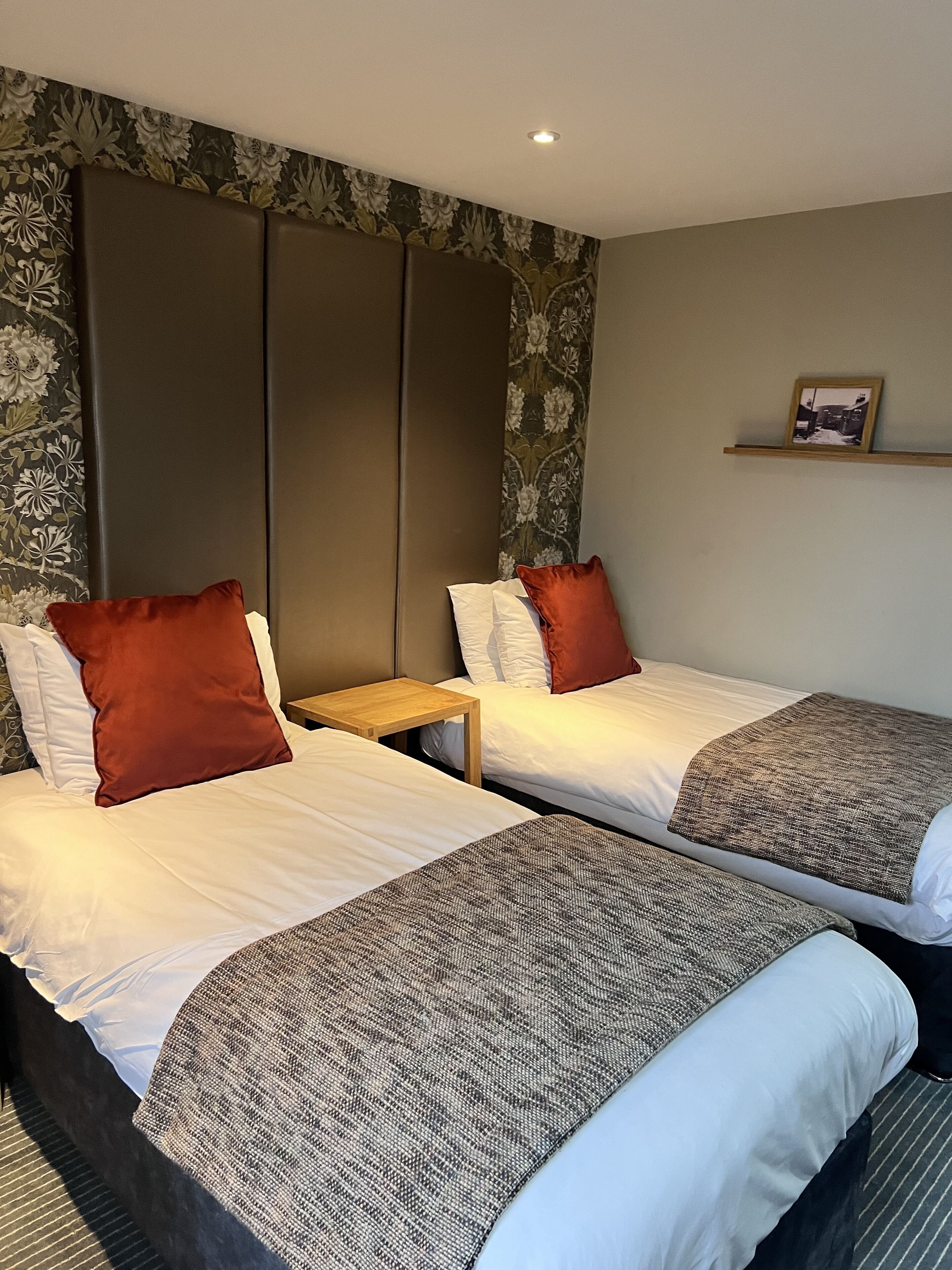 A hotel room with two beds, each with white linens and brown pillows. There is a wooden nightstand between the beds, and a small shelf above the right bed with a framed picture.