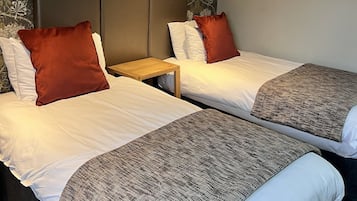 Et hotelværelse med to senge, begge med hvide sengetæpper og brune puder. Der er et trænatbord imellem sengene, og en lille hylde over sengen til højre med et indrammet billede.