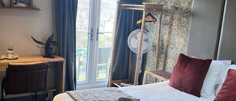 A hotel room with a bed, a desk, a chair, a window with curtains, a wall-mounted fan, and a view of the outdoors.