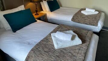 A hotel room with two beds, each with white linens and teal pillows. There is a lamp on one nightstand and a folded towel on each bed.