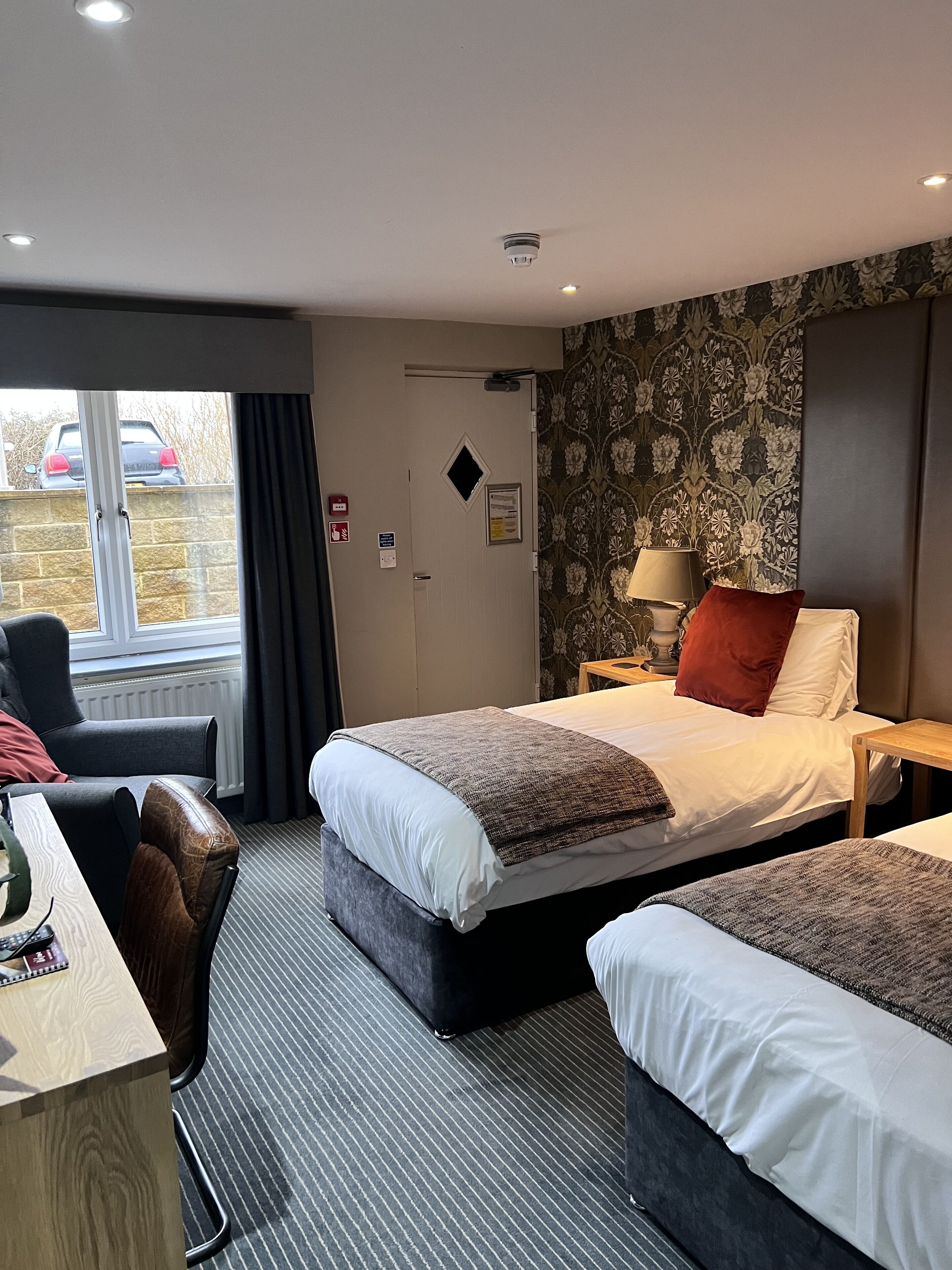 A hotel room with a bed, two bedside tables, a desk, a chair, and a window with a view of a parked car outside.