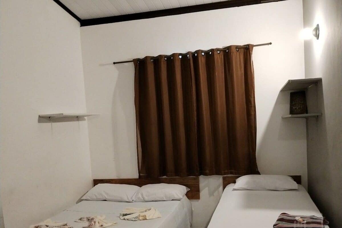 A room with two single beds, a ceiling fan, a window with brown curtains, and a small shelf with a book.