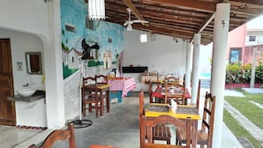 A covered outdoor dining area with wooden tables and chairs, a mural on the wall, and a white sink in the corner. - Pousada Raysol Arraial d'Ajuda (Porto Seguro)