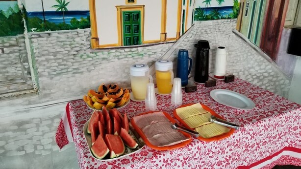 A dining table with a tropical mural, a variety of fruits, and a traditional meal setup. - Pousada Raysol Arraial d'Ajuda (Porto Seguro)