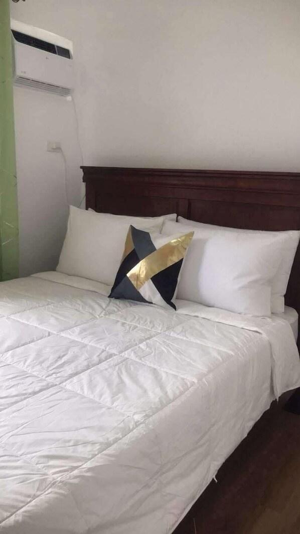 Una cama bien hecha con ropa de cama blanca y dos almohadones decorativos, un cabecero de madera y un equipo de aire acondicionado en la pared.