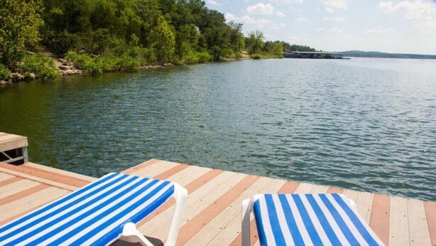 A wooden dock with two striped lounge chairs facing a calm lake, surrounded by dense greenery. - Branson | Cozy Waterfront Retreat | Pool + Lake (Branson)