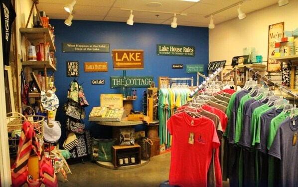Una tienda con varias prendas de vestir en estanterías y repisas, incluyendo camisetas y bolsas. Carteles en la pared señalan diferentes secciones como 'Lake' y 'The Cottage'.