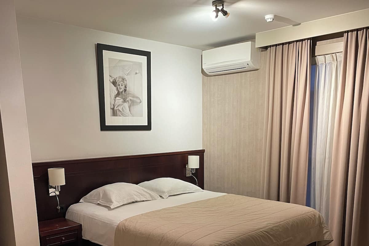 A hotel room with a large bed, a nightstand, a lamp, a framed picture on the wall, an air conditioning unit, and a chair.