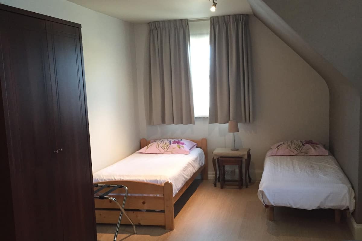 A bedroom with two single beds, a wooden wardrobe, a small bedside table with a lamp, and a window with curtains.