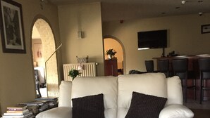A living room with a white sofa, black cushions, a black coffee table, and a patterned rug. There is a bookshelf with books and a small table with a lamp. - Hotel Prelude (Aalter)