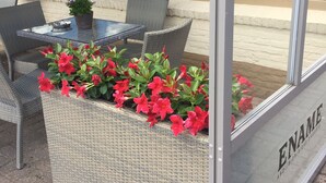 An outdoor seating area with wicker furniture and a glass table, a planter with red flowers, and a view of the street through a glass window. - Hotel Prelude (Aalter)