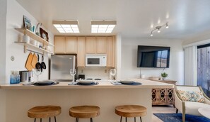 A modern kitchen with a central island, bar stools, wooden cabinets, a microwave, and a flat-screen TV.