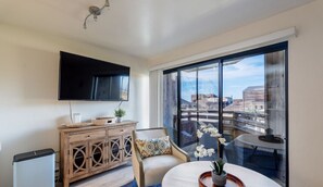 A modern hotel room with a large window, a flat-screen TV, a wooden cabinet, a round table, and a chair.