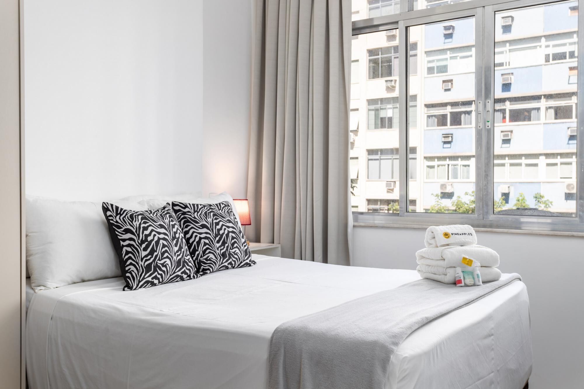 A hotel room with a bed, a window with curtains, and a view of buildings.