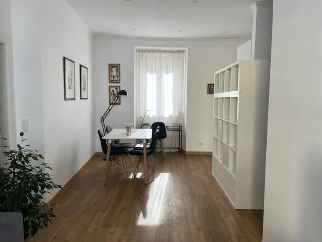 Ein Zimmer mit Holzboden, einem Esstisch mit Stühlen, einer Stehlampe und einem Fenster mit Vorhängen.