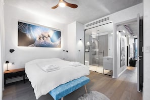 A modern bedroom with a large bed, a bench, a wall-mounted artwork, a glass shower, and a hallway leading to a bathroom. - Top TLV Grand Suites (Tel Aviv)
