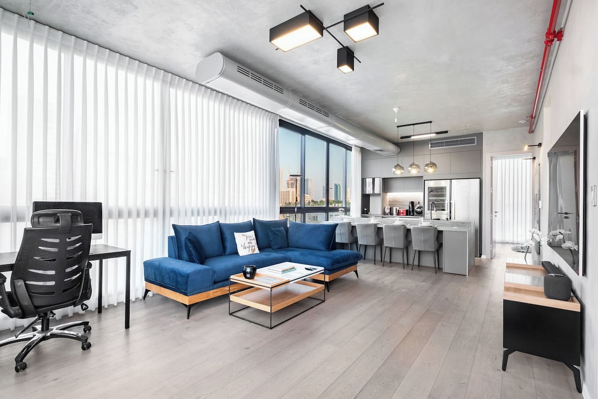 Ein modernes Wohnzimmer mit einem blauen Sofa, einem Couchtisch und einem Schreibtisch mit Stuhl. Es gibt eine Küchenzeile mit einer Bar und ein großes Fenster mit Blick auf die Stadt.
