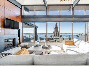 A modern living room with a fireplace, large windows, and a view of the water. - Luxury Muskoka Waterfront Cottage | 5BR | Sauna, EV Charger. Sleep 12 (Port Severn)