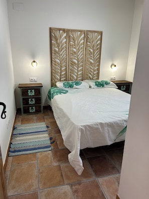 A bedroom with a bed, a nightstand, a decorative headboard, a small dresser, and a wall-mounted light fixture. - Casita en Aldea de Pescadores, en Medio del Pinar y la Playa. Olvídate del Coche (Monte Gordo)