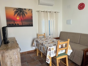 A cozy living room with a sofa, a dining table with chairs, a TV, and a painting of a sunset. - Casita en Aldea de Pescadores, en Medio del Pinar y la Playa. Olvídate del Coche (Monte Gordo)