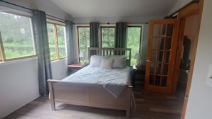 Une chambre à coucher avec un lit, une table de chevet, une lampe, une porte en bois et de grandes fenêtres avec des rideaux.
