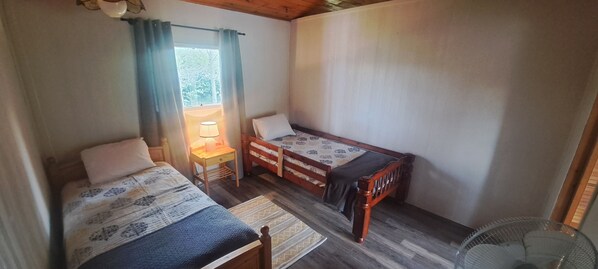 Une petite chambre agréable, dotée de deux lits simples, d’une table de chevet en bois, d’une table avec une lampe et d’une fenêtre avec des rideaux.