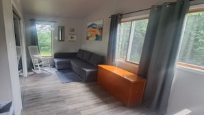 Un espace de vie compact comprenant un canapé gris, un banc en bois, un fauteuil à bascule et une petite table. Une fenêtre avec des rideaux laisse entrer la lumière naturelle et offre une vue sur la verdure à l’extérieur.