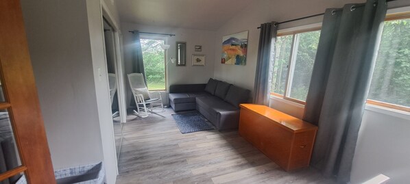 A compact living space with a gray sofa, a wooden bench, a rocking chair, and a small table. A window with curtains allows natural light and views of greenery outside.