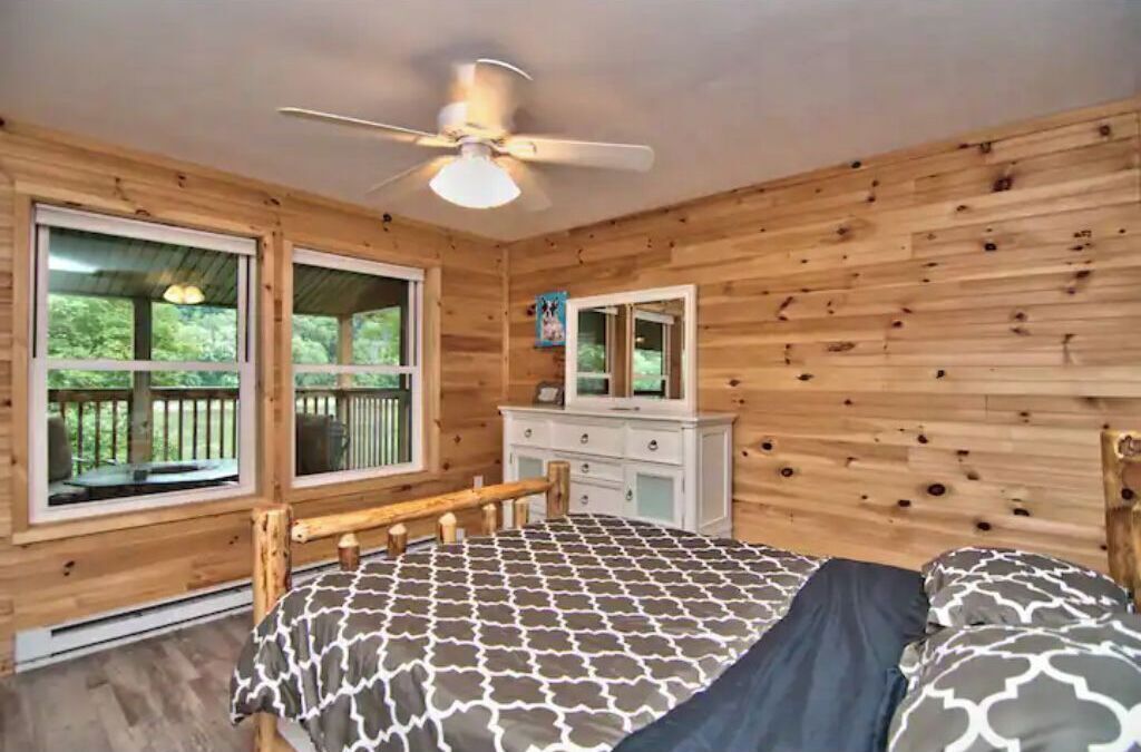 A wooden cabin bedroom with a bed, dresser, and large windows.