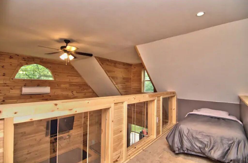 A bedroom with a sloped ceiling, wooden walls, a ceiling fan, a window, and a bed.