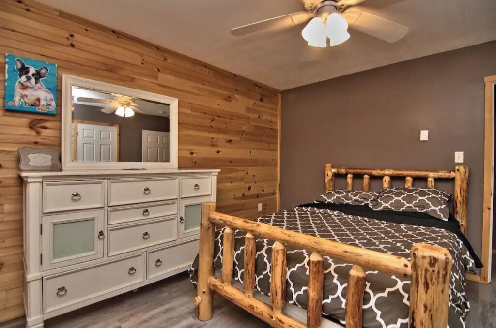 Une chambre à coucher avec un lit en bois, une commode blanche dotée de nombreux tiroirs, un ventilateur de plafond et un grand miroir placé au-dessus de la commode.