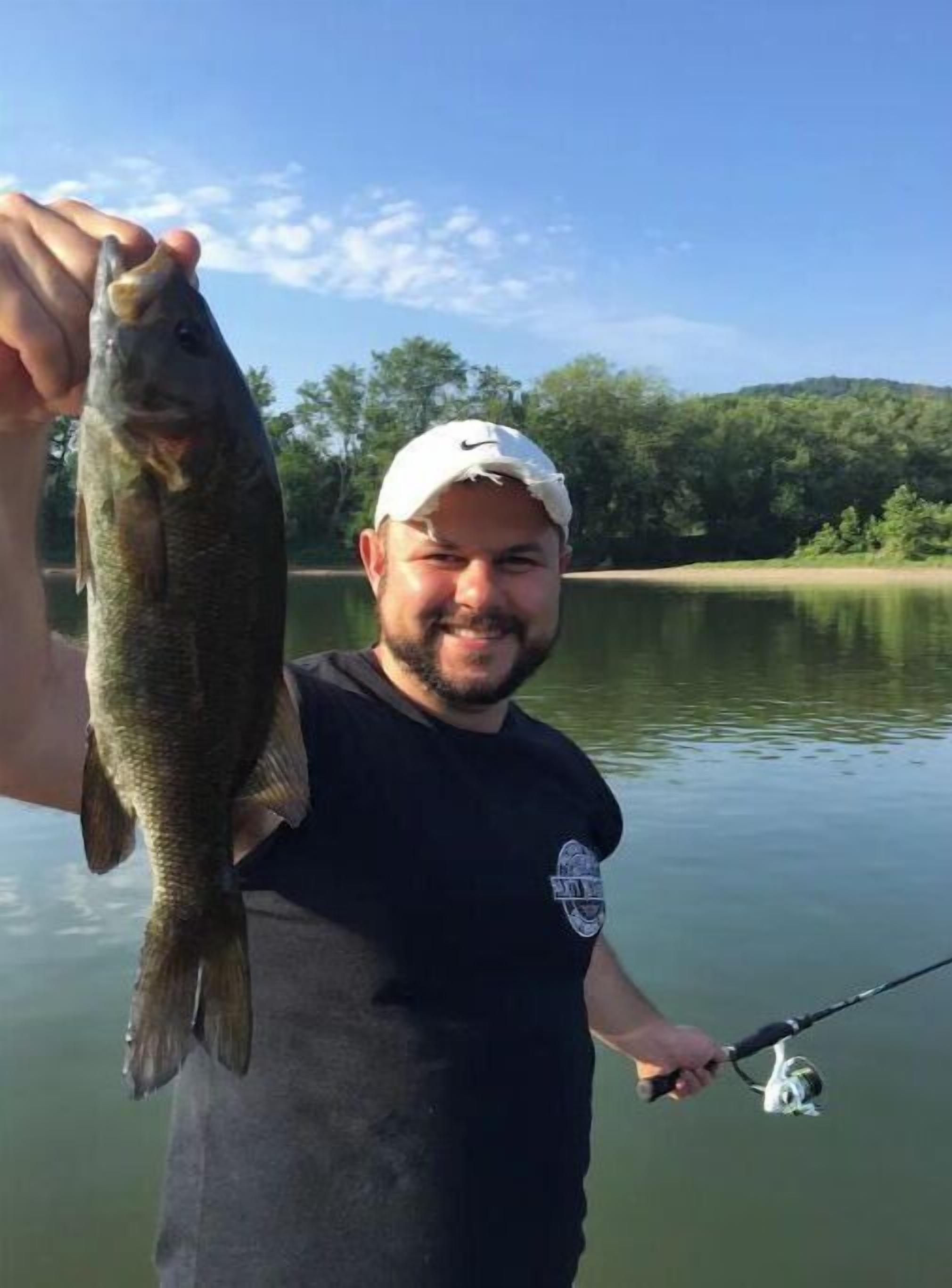 A person holding a fish, standing by a calm lake with trees in the background.