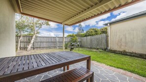 An outdoor patio area with a wooden table and benches, a stone pathway, and a wooden fence in the background.