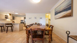 Uma sala de jantar com mesa e cadeiras de madeira, uma cozinha ao fundo, e um quadro na parede.