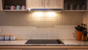A kitchen with a black induction cooktop, a tiled backsplash, and a shelf with mugs and utensils.