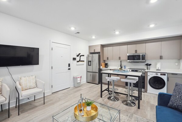 A modern kitchen with a microwave, refrigerator, and washing machine. There is a dining area with a table and bar stools. A flat-screen TV is mounted on the wall.