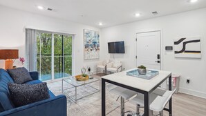 Un salon moderne avec un canapé bleu, une table basse en verre et une table à manger avec des chaises. Une grande fenêtre offre une vue sur la verdure à l’extérieur.