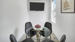 A modern dining area with a glass table, two chairs, a vase of flowers, a flat-screen TV mounted on the wall, and a framed picture on the wall.