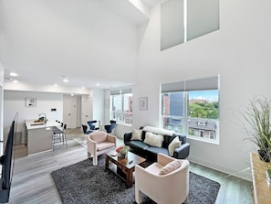 Un salon moderne avec un canapé noir, des fauteuils blancs et une table basse. On y trouve également un coin repas avec une table et des chaises blanches, et une cuisine à l’arrière-plan.