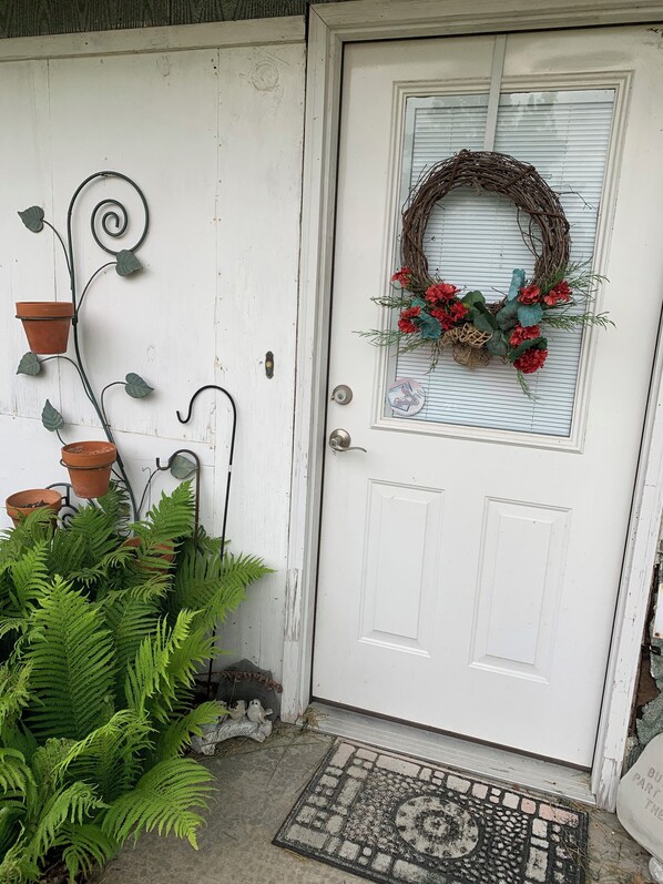 Une porte blanche ornée d’une guirlande, avec des plantes en pot et un paillasson.