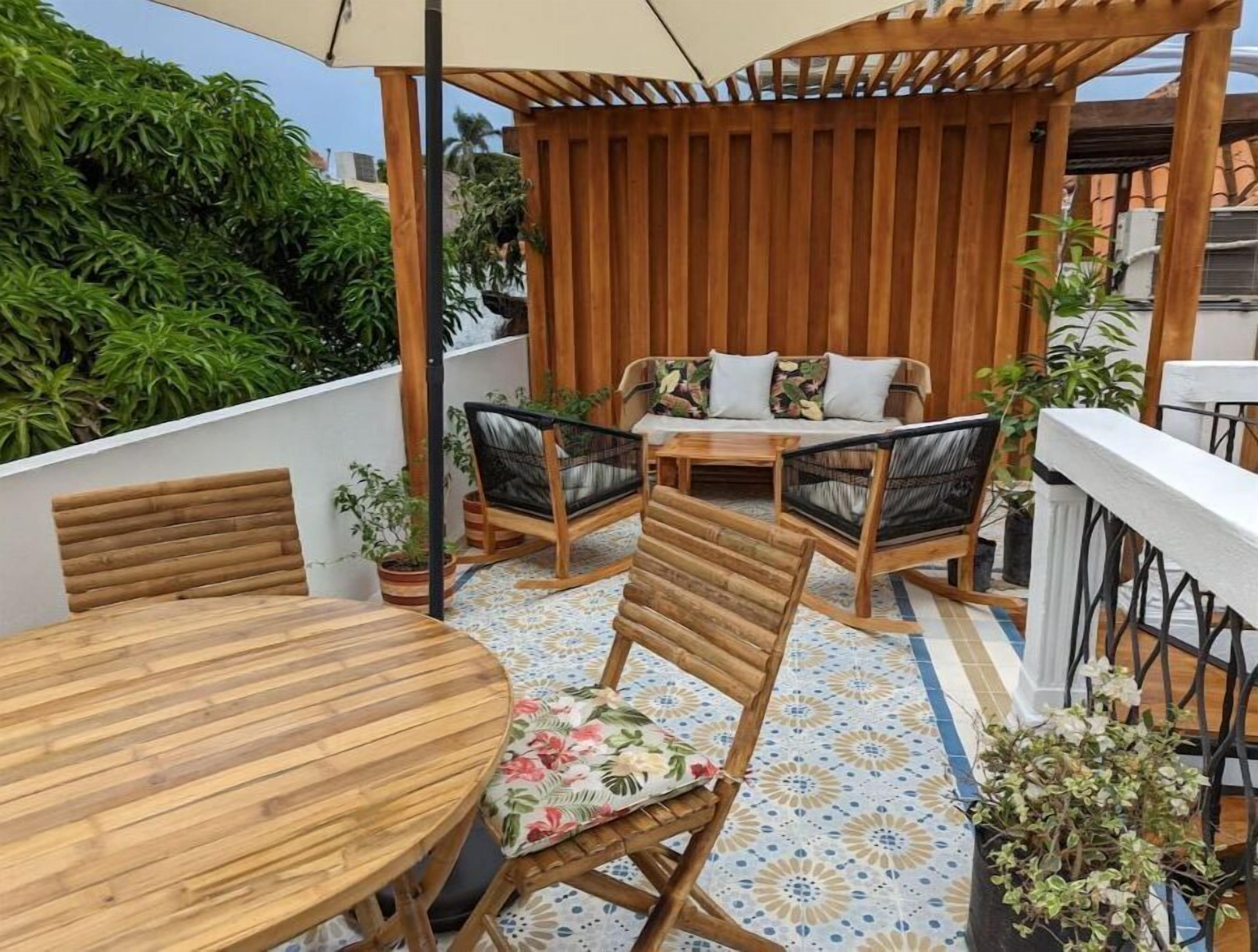 Et udendørs opholdsområde i træ med et rundt bord, stole, en sofa og krukker med planter.