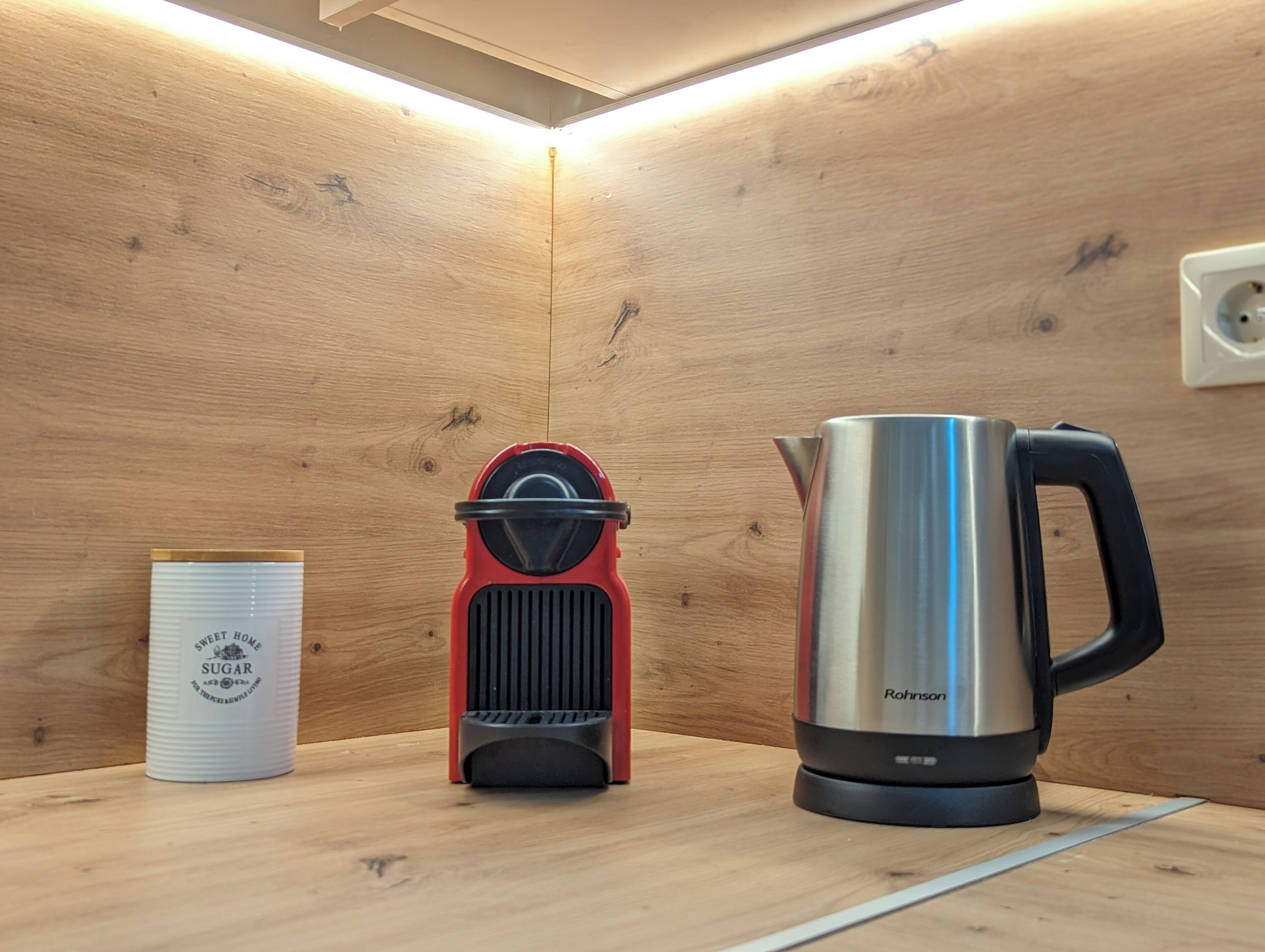 Una kitchenette con una cafetera de acero inoxidable, una tetera roja eléctrica y un recipiente blanco etiquetado 'SWEET HOMES SUGAR'.