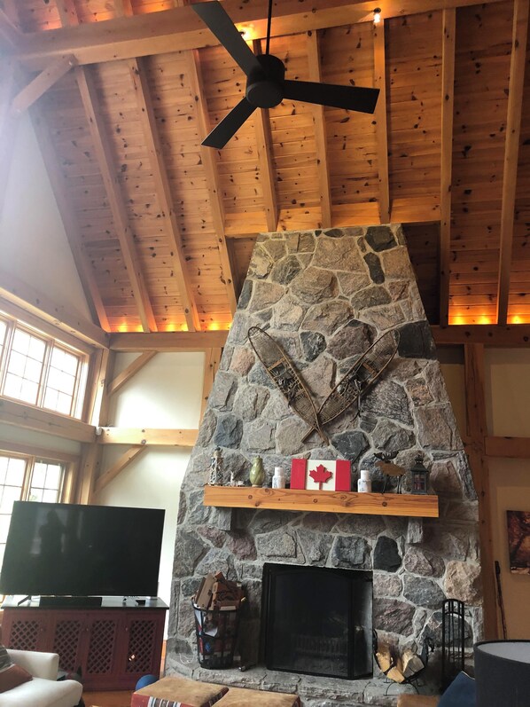 A cozy living room with a stone fireplace, wooden beams, a ceiling fan, and a mounted television.