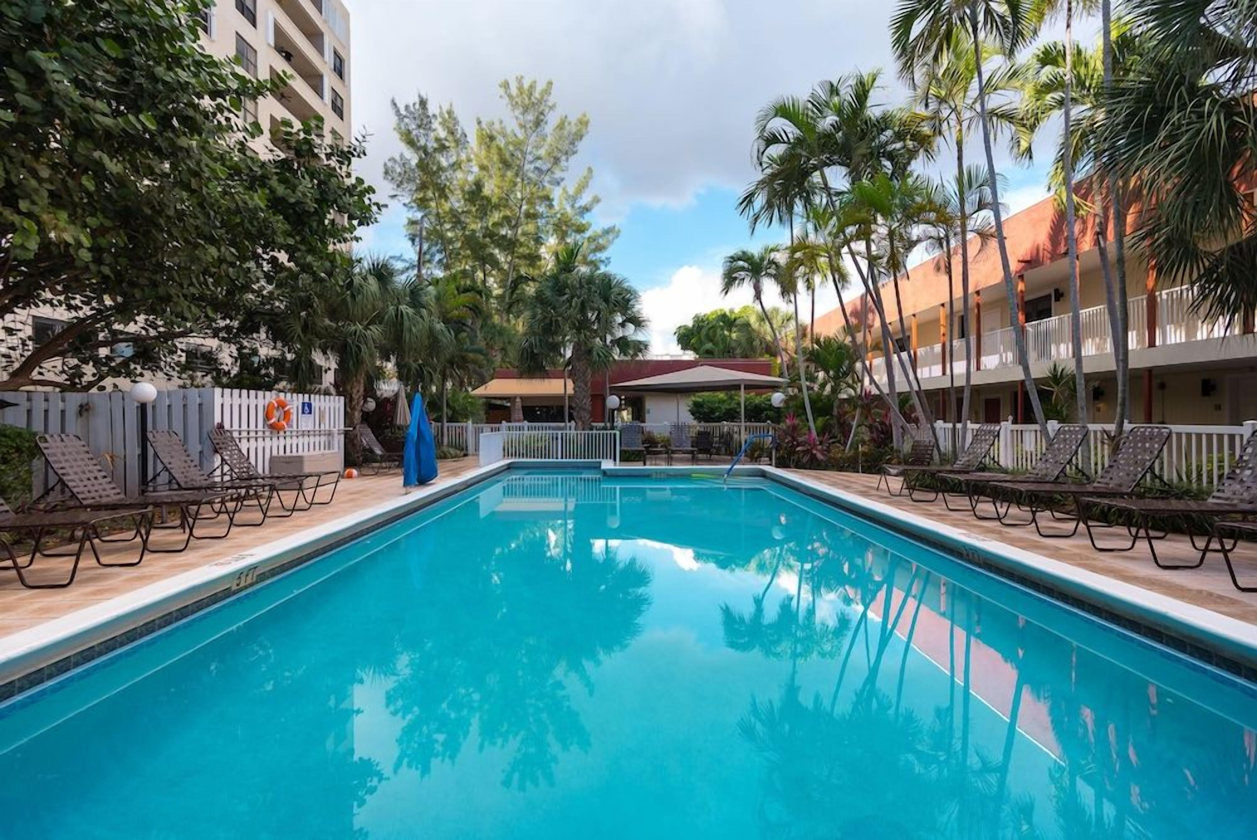A swimming pool with lounge chairs, surrounded by palm trees and a building with balconies.