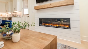 Ett modernt vardagsrum med en öppen spis, en platt-TV som hänger ovanför, och ett köksområde i bakgrunden.