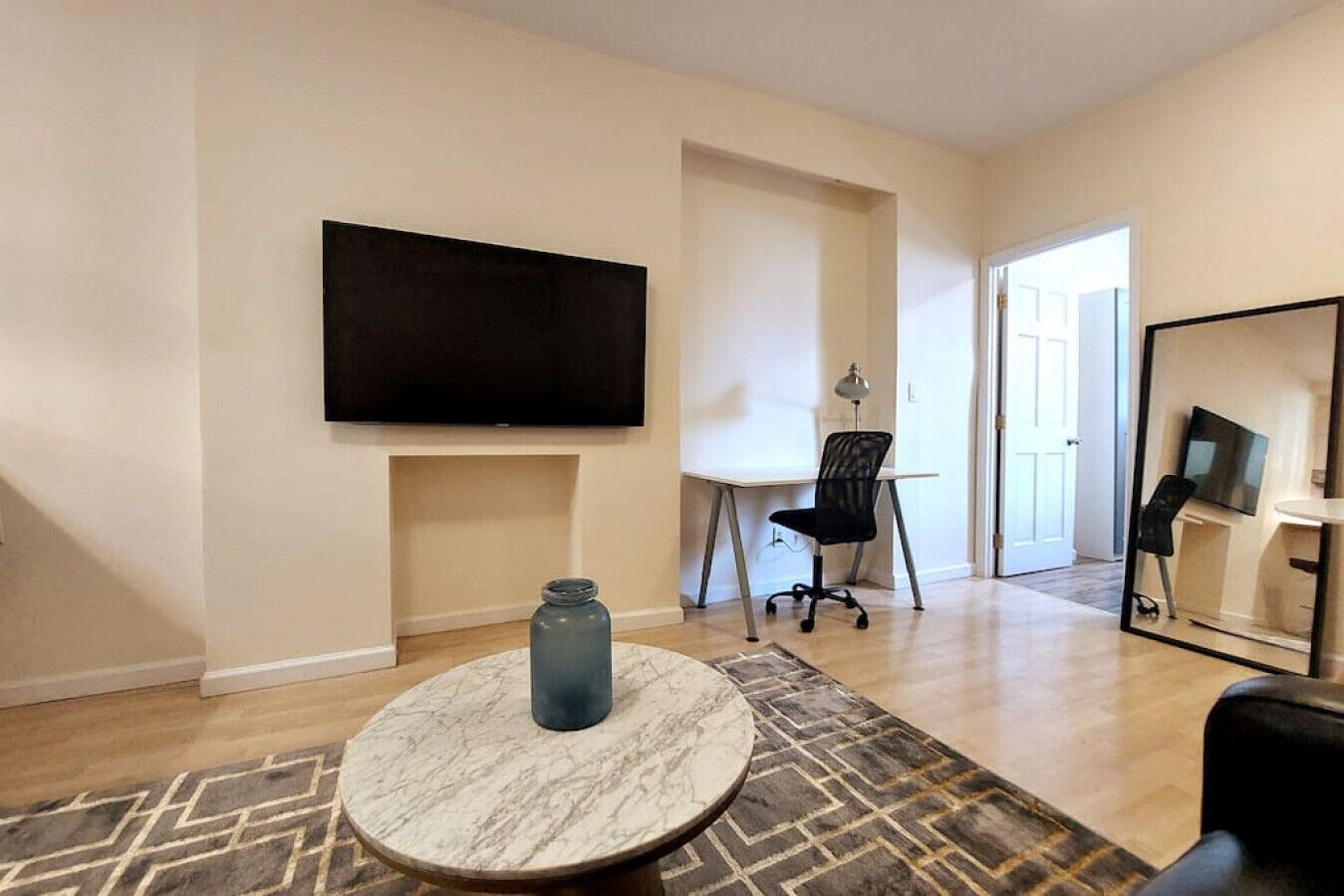 Ett modernt vardagsrum med en platt-TV, ett runt bord i marmor, en svart kontorsstol och en stor spegel.